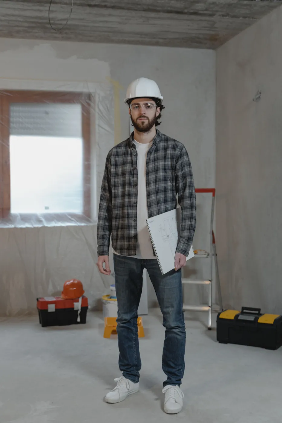 Contractor in a hard hat and safety glasses holding blueprints in a room under renovation