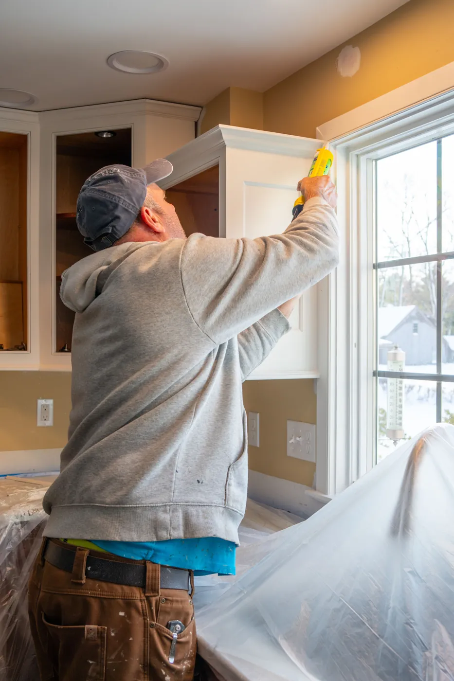 Contractor in a ball cap caulking the trim of a white kitchen cabinet next to a window